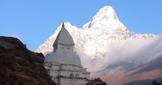 Aerial drone shot capturing snow-capped Mount Ama Dablam rising above a cloudy Himalayan landscape with Buddhist stupa monuments and engraved stone inscriptions in the Everest region of Nepal.