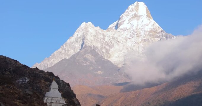 An aerial pull-in shot toward a white Buddhist stupa and engraved stone inscriptions with Mount Ama Dablam rising above drifting clouds under a clear blue sky in the Everest region of Nepal.
