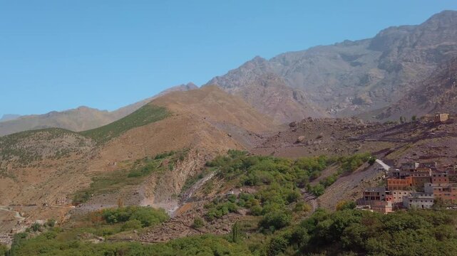 Panoramic view of a traditional Berber village perched on a rocky hillside in Imlil Valley, Morocco, surrounded by rugged slopes and green trees beneath the dramatic peaks of the High Atlas Mountains.