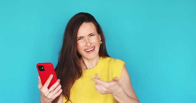 Aggressive and annoyed young woman shouting at smartphone screen, gesturing with anger and frustration, feeling outraged by message or phone glitch, standing against blue background in studio
