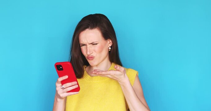 Disgusted young woman looking at smartphone screen with aversion and confusion, showing hand gesture of dislike, frustrated by bad social media content or message, standing on blue background
