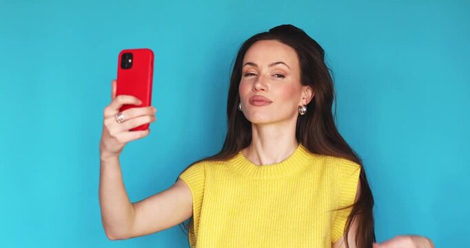 Attractive young woman with long brunette hair wearing yellow knitted vest posing with red smartphone making selfies and video stories smiling making duck face isolated blue background copy space