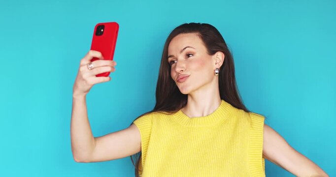 Attractive young woman with long brunette hair holding red smartphone in hand on isolated blue background looking at screen making selfies posing and checking hairstyle smiling making duck face and fi