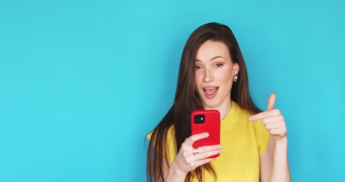 Attractive young woman with long brunette hair holding red smartphone in hands laughing pointing away making video stories or selfie isolated blue background copy space content creator portrait