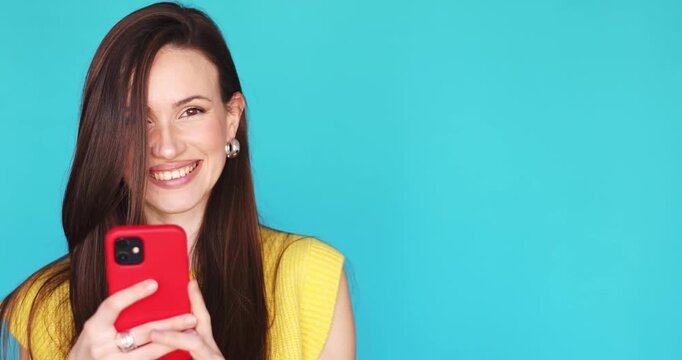 Cheerful young brunette woman holding red smartphone and pointing thumb at copy space while laughing and looking at camera isolated on blue background influencer sharing mobile app or social media new