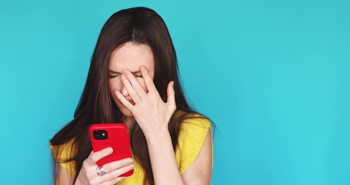 Upset young woman looking at her smartphone and making a facepalm gesture with a disappointed facial expression while standing against a blue background feeling sad and stressed