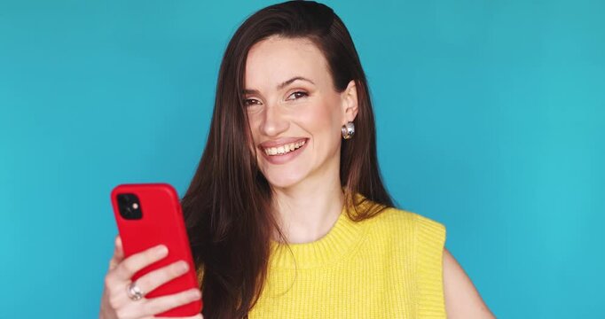Happy young woman laughing and looking at smartphone screen with joy. Cheerful female user in yellow sweater holding red phone and expressing positive emotions on vibrant blue studio background.