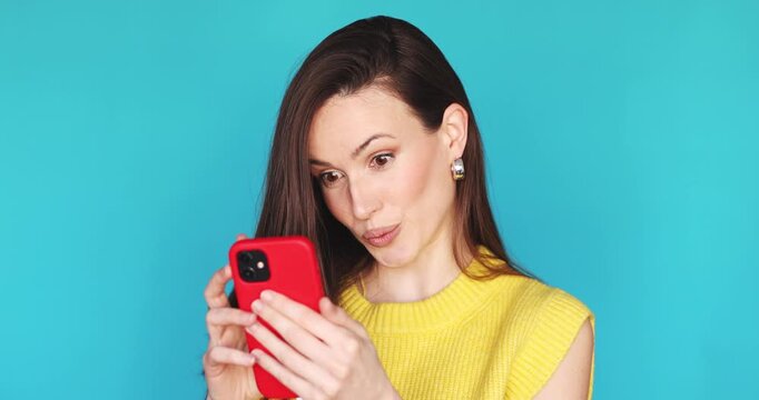 Young woman in yellow sweater using red smartphone and showing different emotions. Surprised female user looking at screen with wow expression and smiling on vibrant blue studio background.