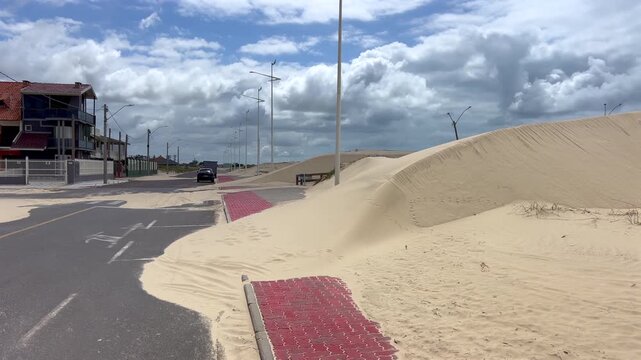 Sand dunes advancing over sidewalk bike lane and paved street in a coastal urban area under cloudy sky.