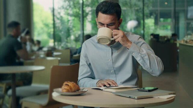 Sleepy Indian business man yawning feeling tired drinking coffee writing notes ideas in cafe working distant in morning overworked Arabian businessman write drink guy male lack energy problem insomnia