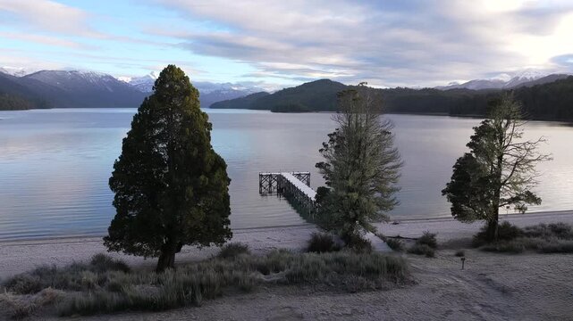 Drone advancing toward wooden pier on Lago Espejo with snowy Andes mountains in Patagonia.