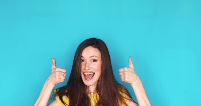 Excited young woman pointing fingers up at copy space and showing double thumbs up isolated on blue background, happy female brunette recommending product or service, wow emotion and approval