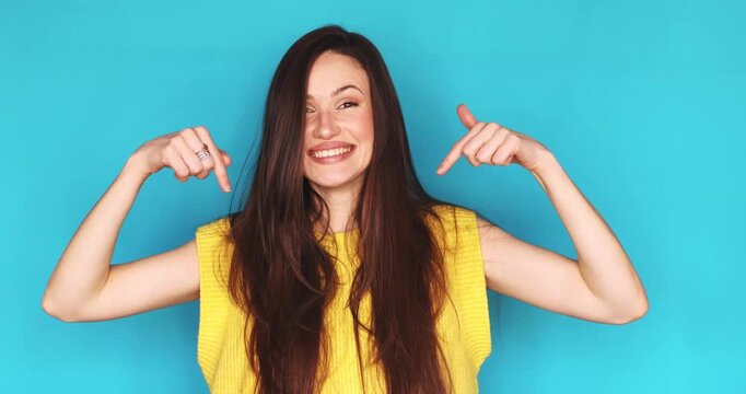 Cheerful young woman pointing fingers down at copy space and laughing sincerely isolated on blue background, happy female brunette recommending link or product, expressive emotions and joy