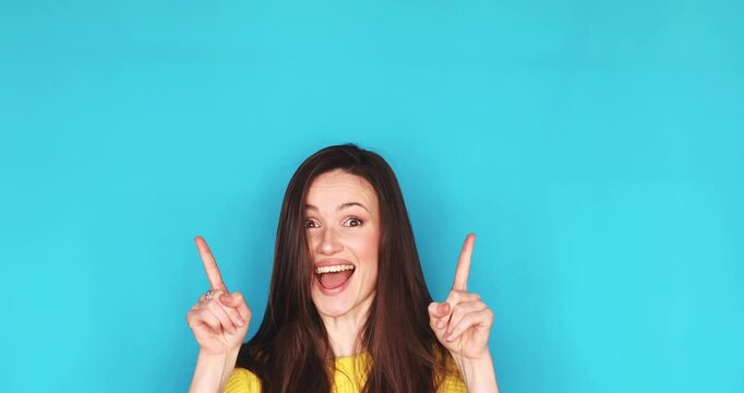 Surprised and happy young brunette woman pointing fingers up at copy space above her head isolated on blue background, excited female showing amazing offer or big news with wow face expression