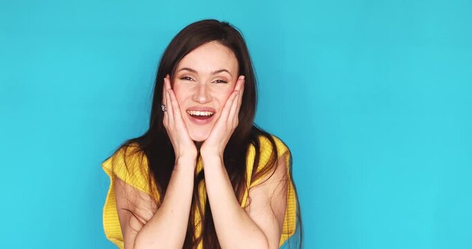 Amazed and shocked young brunette woman looking at camera with open mouth and hands on cheeks isolated on blue background, happy female feeling surprised and excited, wow face expression