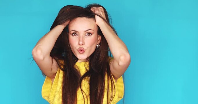 Shocked and stressed young brunette woman grabbing hair and looking around in panic isolated on blue background, surprised female feeling overwhelmed or confused, expressive facial reaction