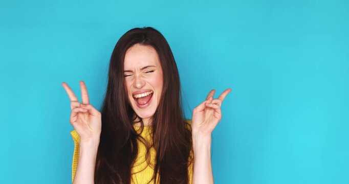 Cheerful young woman with long dark brown hair smiling and showing victory peace sign with both hands on turquoise background wearing yellow knitted top, expressing pure joy, happiness and positivity.