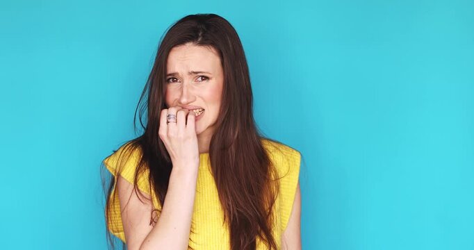 Nervous young woman biting her nails and looking away with worried face expression, feeling anxious and stressed, standing isolated on blue background. Concept of anxiety, fear, and panic attack