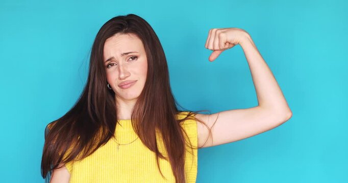 Funny young woman showing her weak biceps and pointing at it with disappointed face expression, feeling unconfident about her muscles, standing isolated on blue background. Concept of fitness failure