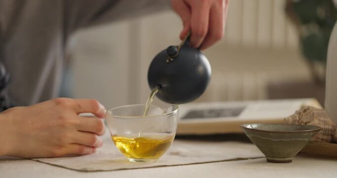 Hands delicately pour fragrant green tea into a glass cup, inviting tranquility and warmth in a homey atmosphere