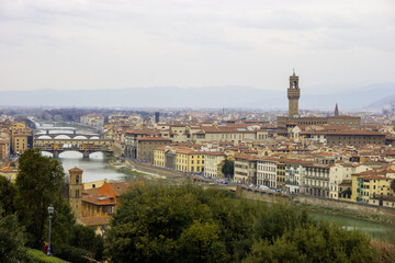 Obraz premium A panoramic view of Florence, Italy, showcasing the Arno River, Ponte Vecchio, and the Palazzo Vecchio tower under a cloudy sky.