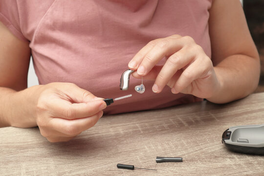 Woman cleaning a digital hearing aid with a small brush tool on a wooden table, hearing aid maintenance and care