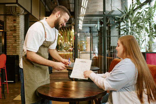 Biracial young adult man wearing apron standing and smiling, while taking order from Caucasian young girl sitting at table holding menu in modern restaurant interior