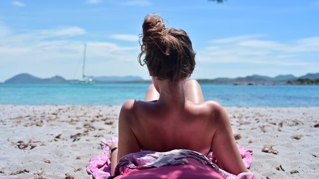 Rear view of an attractive woman sitting on a beautiful sandy beach looking at the sea. Relaxing summer scene with ocean horizon, travel, vacation and freedom concept.