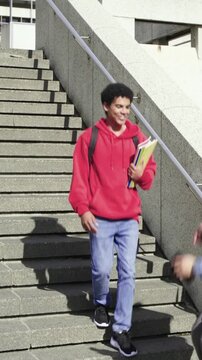 Vertical video: Walking teen red hood holding folders campus steps friend approaching, swapping bag