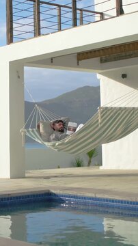 Vertical video: Panning camera revealing Indian man reading book in striped hammock, wearing hoodie
