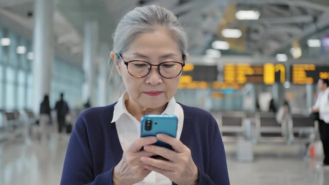 Senior woman using smartphone, checking email or social media, in modern airport terminal, showing technology adoption.