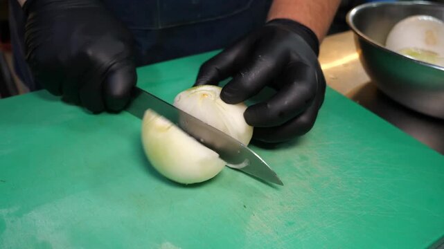 A chef wearing black gloves and a denim apron meticulously dices white onions on a green cutting board in a professional kitchen. The chef uses precise cutting techniques with a sharp chef's knife, tr