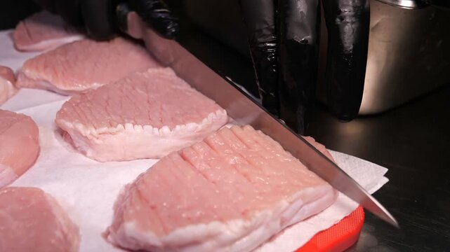 A chef scores and tenderizes raw pork chops with a knife and specialized tool on a cutting board in a professional kitchen.