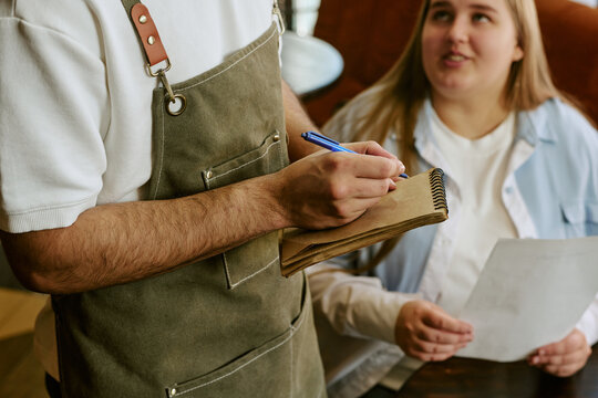 Caucasian young adult man wearing apron taking order from Caucasian young adult woman sitting at table holding menu, focusing on interaction between server and customer in restaurant setting