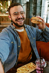 Vertical portrait of Young adult biracial man smiling at camera while holding burger in one hand,...
