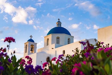 Obraz premium A Greek Orthodox church with blue domes and bell towers is framed by vibrant purple flowers, set against a bright sky.