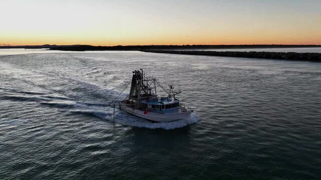 Closing in on a working prawn trawler, the drone captures it leaving the river at sunset. Soft golden light reflects off the water as the vessel heads out toward the open ocean and evening swells
