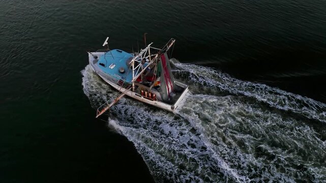 Drone glides around a departing prawn trawler at late sunset, its wake rippling through the river mouth as it heads toward the open ocean. Golden light fades on the horizon and gentle waves r