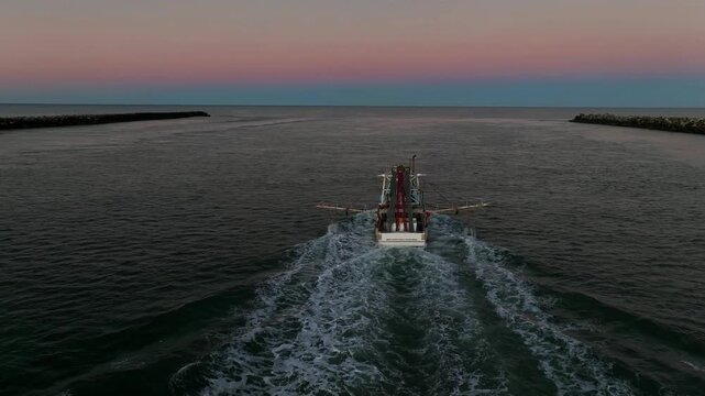 Flying low, the drone approaches a prawn trawler leaving the river at dusk. The boat pushes toward the open ocean as the last orange glow of sunset reflects across calm water and small waves.