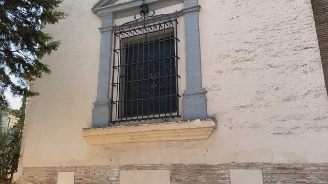 shot of the rear facade of a small chapel. A large window with old wrought iron bars is framed by decorative columns. As the camera rises, the triangular roof and a round oculus appear.