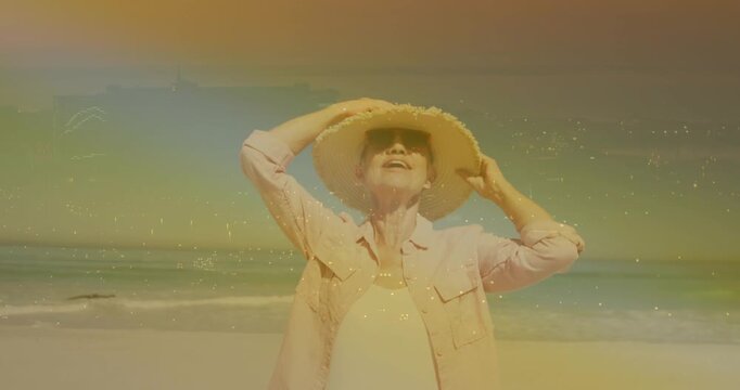 Senior woman feeling warm sun, holding hat, adjusting brim on beach, causing sparkles for travel