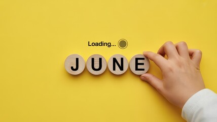 Naklejka premium Hand placing a wooden block with the letter 'E' to complete the word 'JUNE' on a yellow background, with loading text.