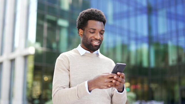 Smiling african american businessman using a mobile phone while standing on street near a modern office building. Happy worker in casual sweater checks smartphone, chats online, browses web. Close up