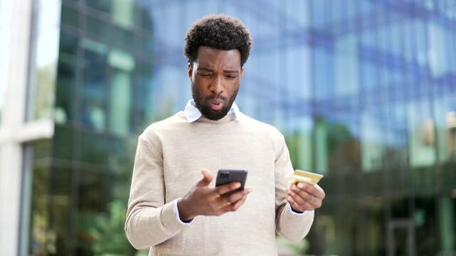 Frustrated african american businessman checks his credit card on phone standing on the street near office building. Shocked worker faced fraud, money was stolen from his account, victim of deception