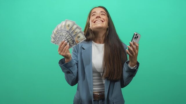 Woman holding a fanned stack of dollars and smartphone, smiling and raising hands in studio; financial success joy.