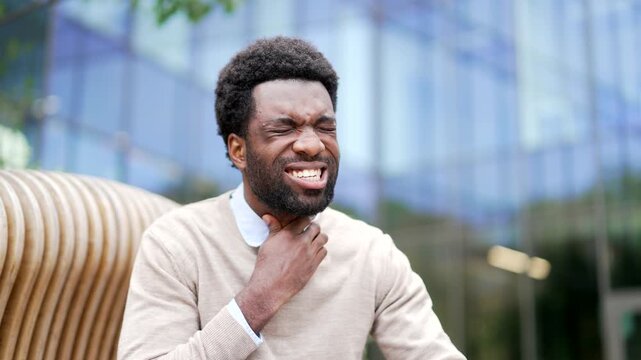 Sick African American businessman suffering from a sore throat sitting on bench on street near a business office building. Unhealthy man coughs, has cold, virus or flu, massaging a sore spot. Close up