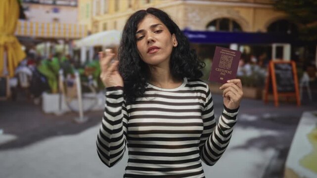 Woman holding passport and finger on chin wearing striped sweater on street cafe terrace; curiosity travel planning decision.