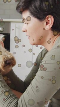 Vertical video: Woman leaning into dog, cupping head, petcare bonding in kitchen via bubble overlay