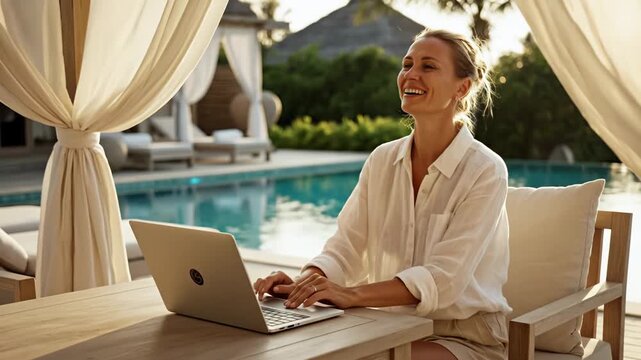 Caucasian woman working on laptop outdoors by swimming pool, smiling and enjoying the serene environment with sunlit curtains and tropical greenery in the background