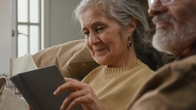 Close-up view of smiling elderly lady with grey hair reading book and looking at positive husband nodding his head while sitting next to her